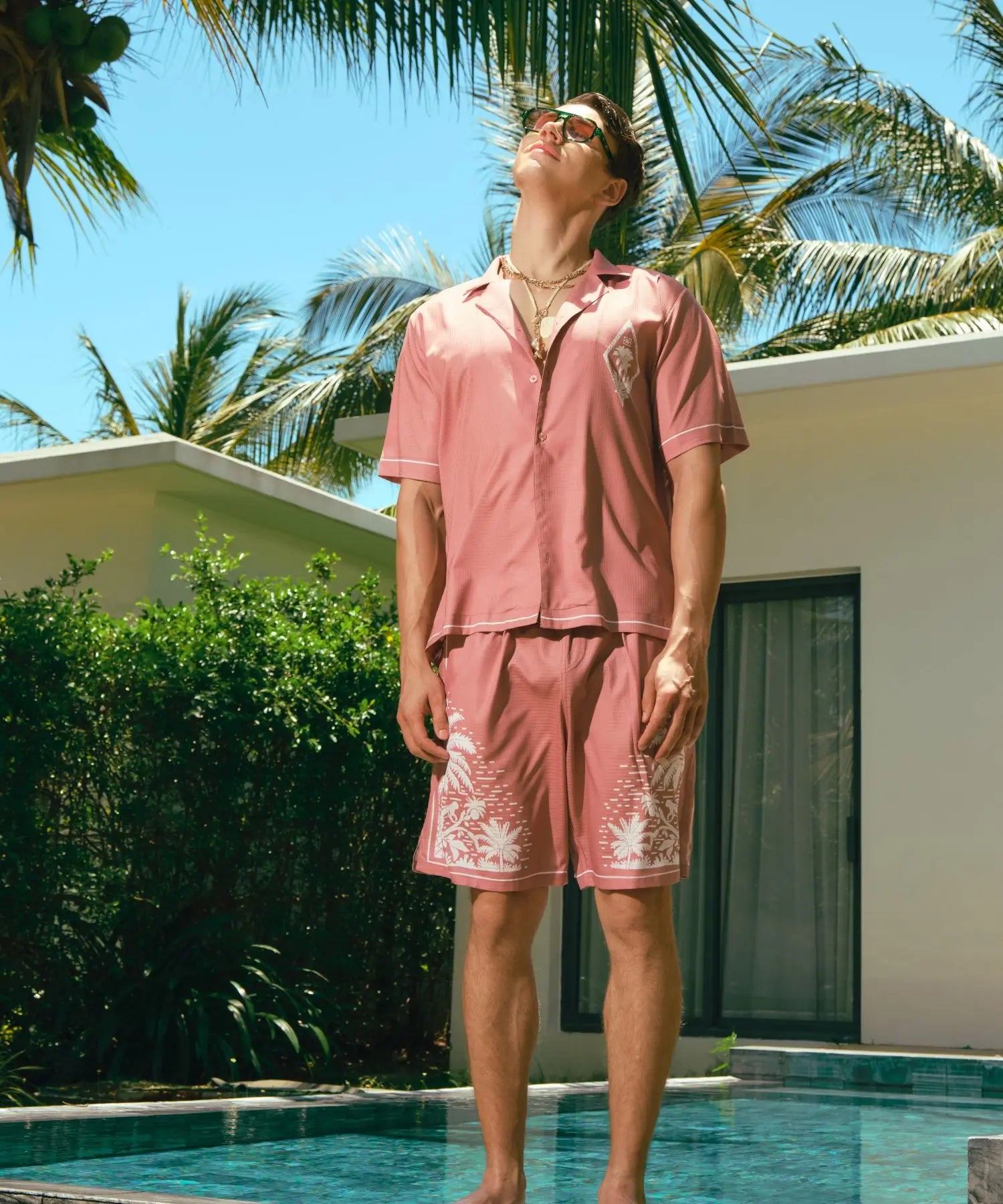 Person wearing a pink outfit with white embroidery by a poolside with palm trees in the background