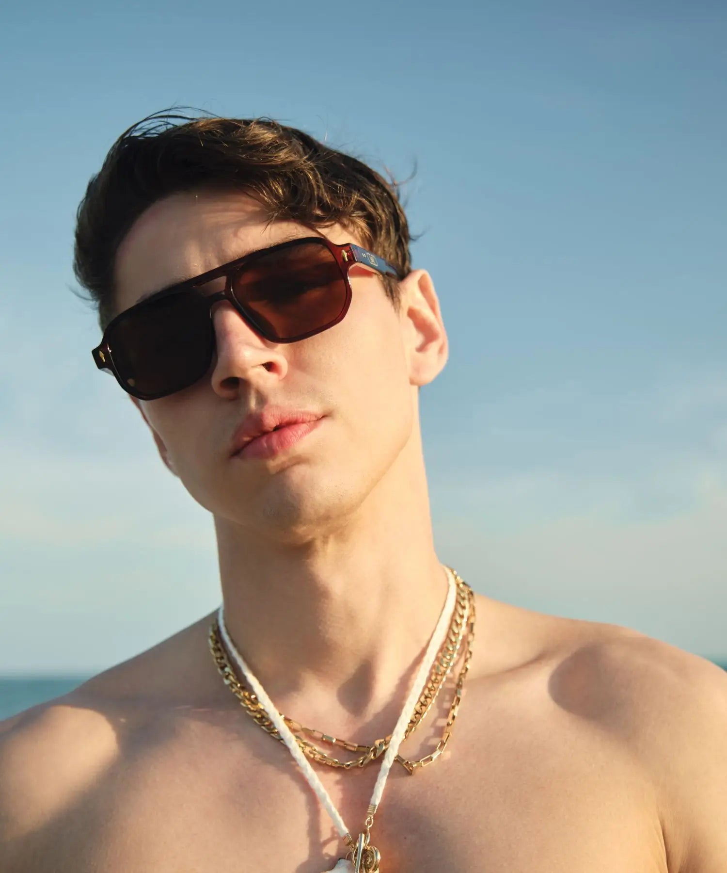 Man wearing sunglasses and gold necklaces against a clear blue sky
