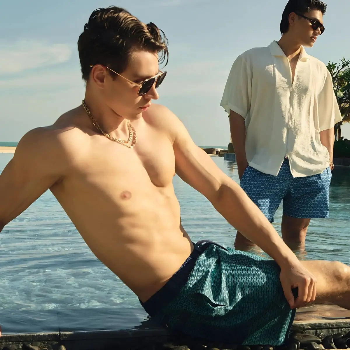 Man sitting by a pool wearing sunglasses and a necklace, with another man standing in the background.