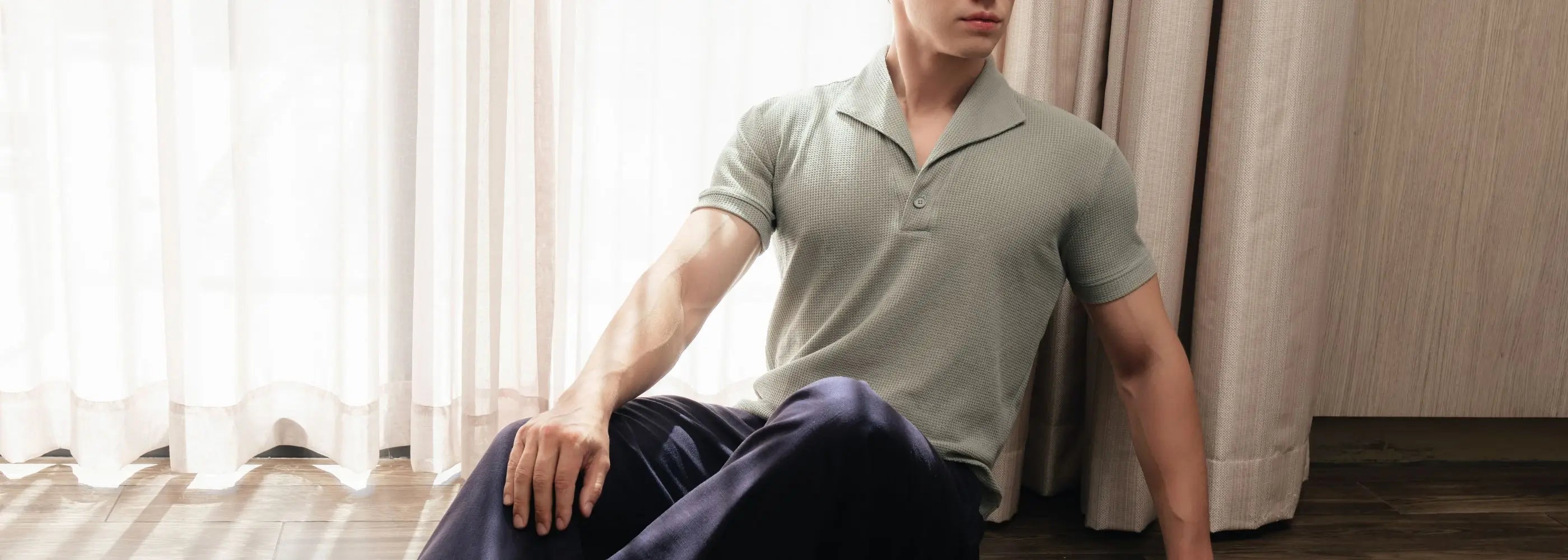 Man sitting on the floor wearing a light green polo shirt and dark pants in a room with white curtains.