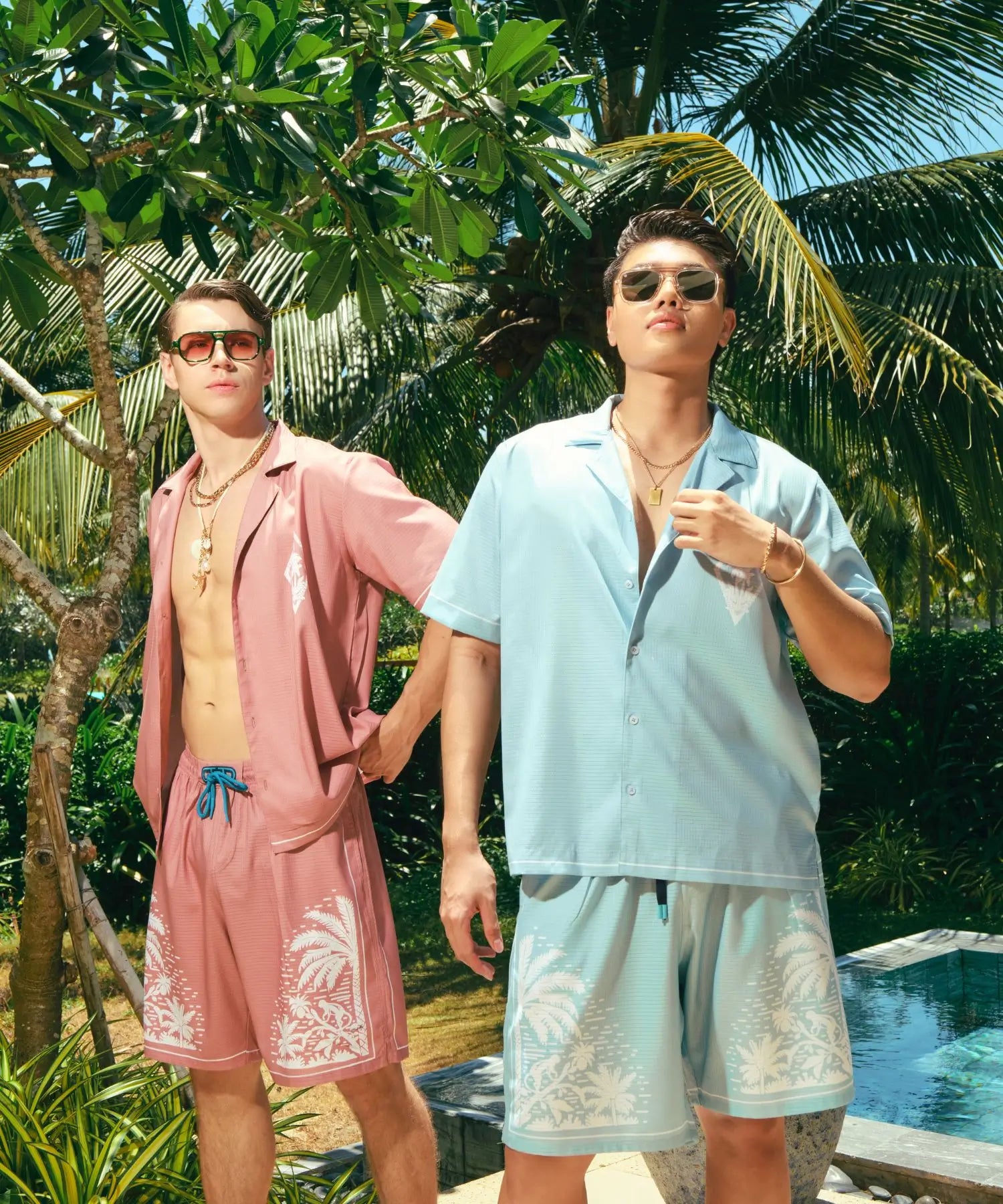 Two men in tropical attire standing in a lush garden with palm trees and a pool.