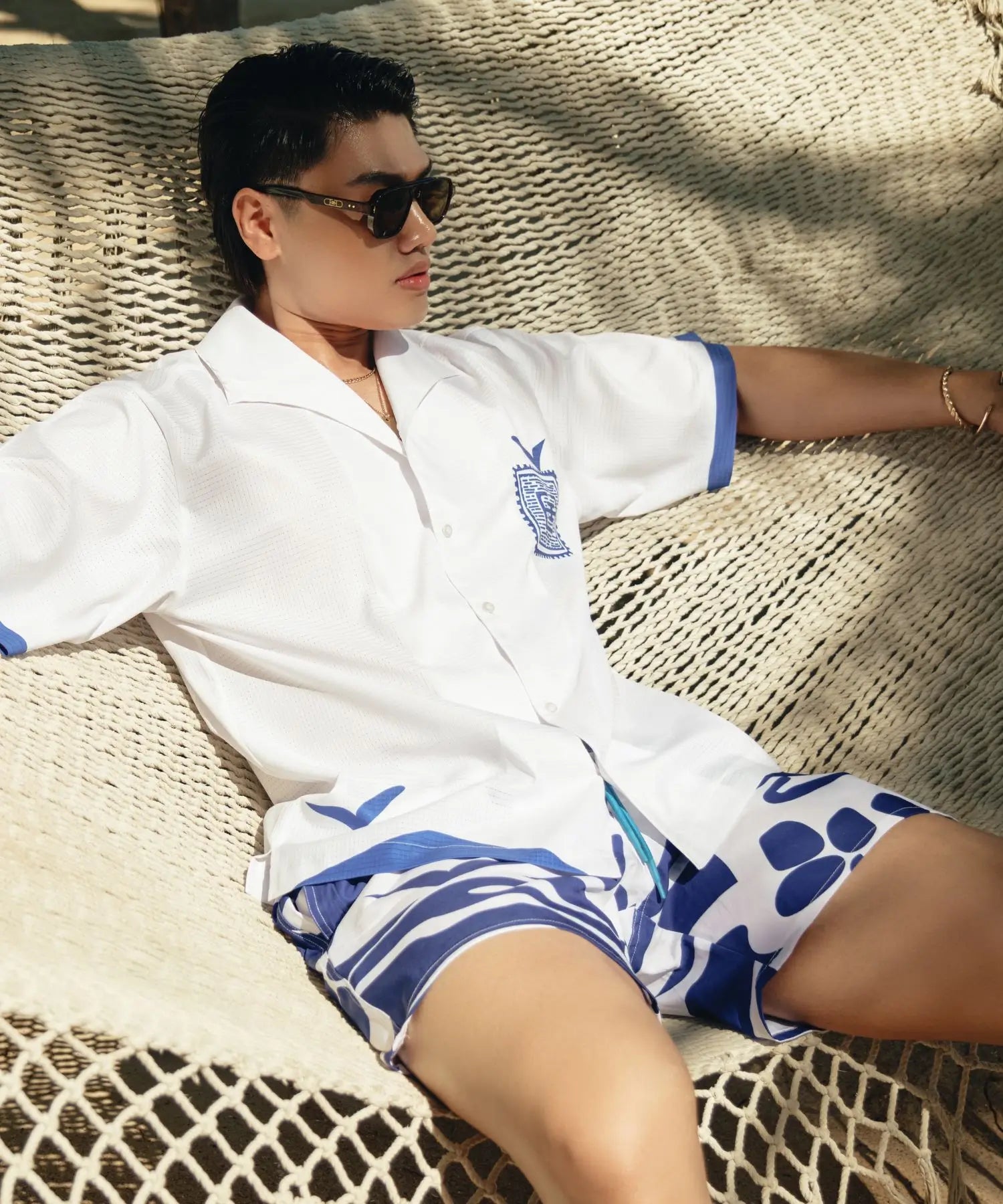 Person wearing a white shirt with blue accents and shorts, sitting on a woven surface.
