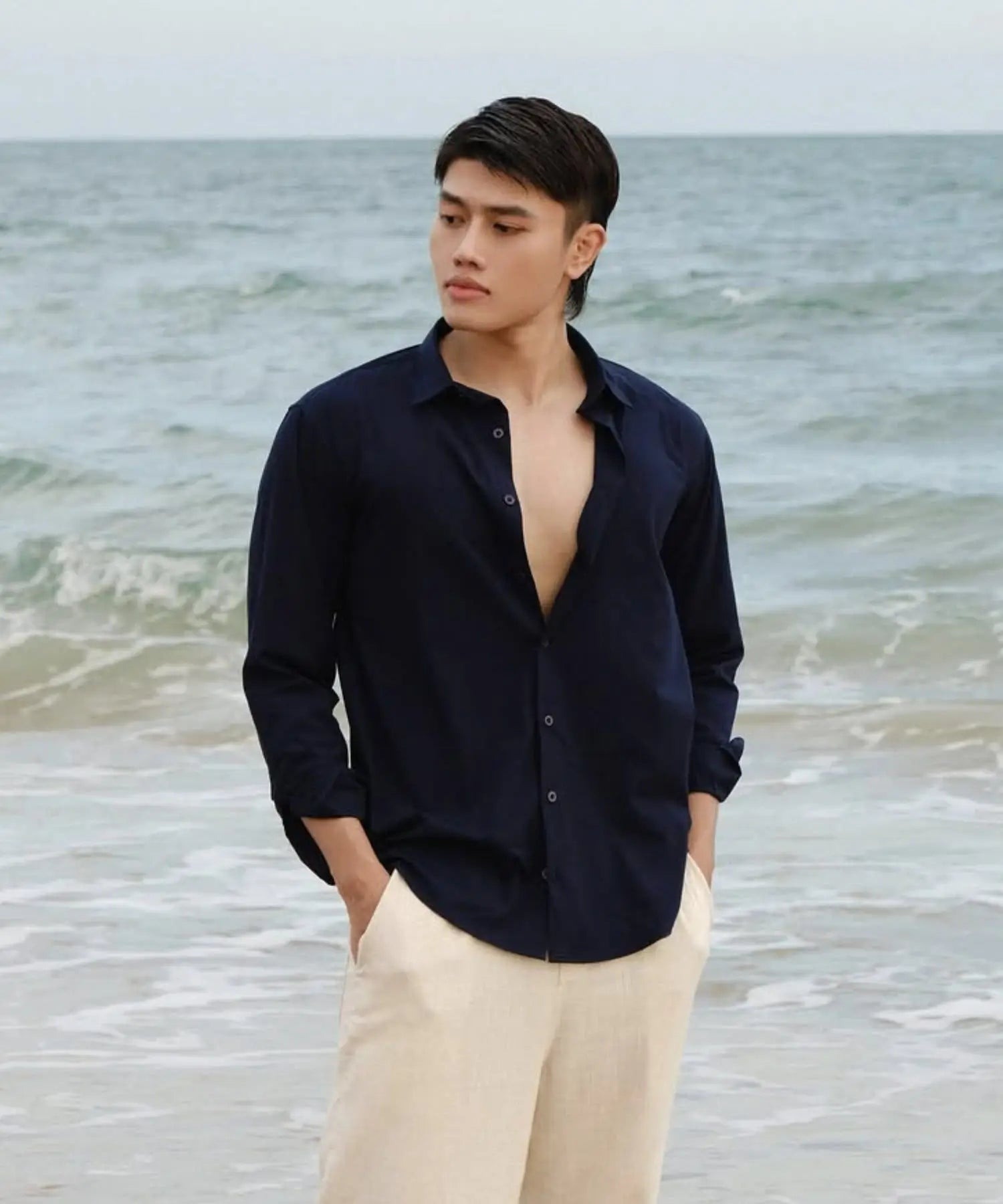 Man wearing a navy shirt with the top button undone, standing on a beach.