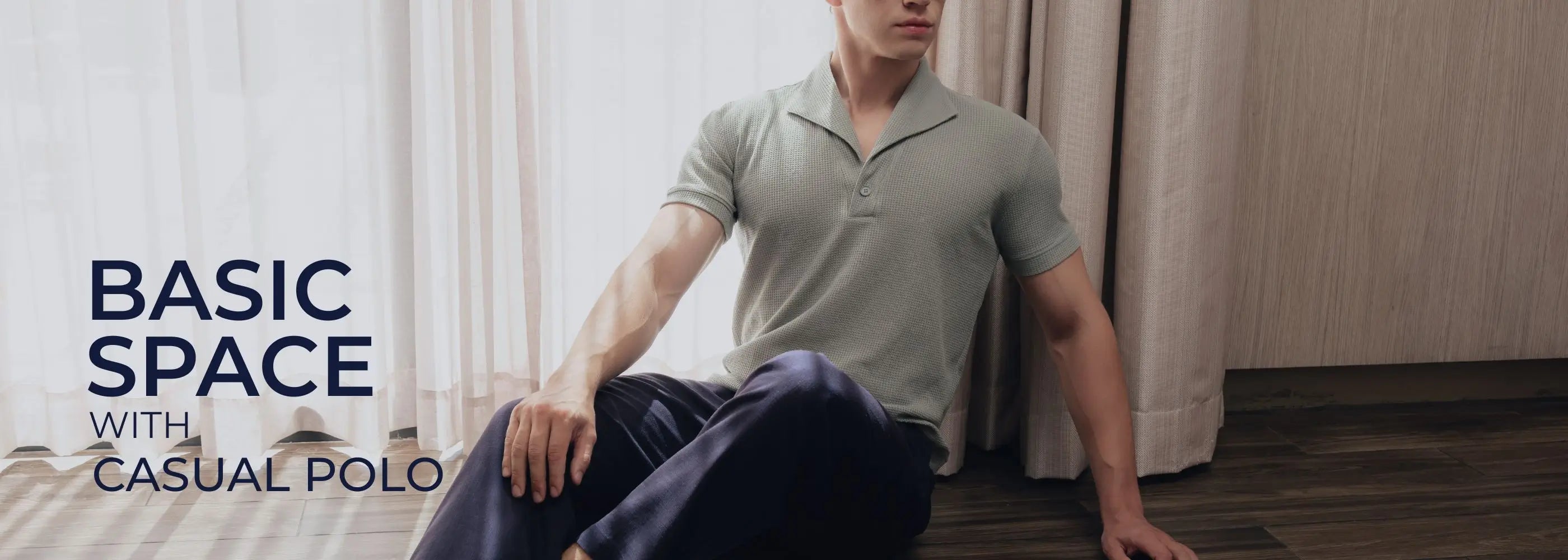 Man sitting on the floor wearing a light gray polo shirt and dark pants in a minimalistic room.